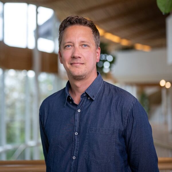 A man in a blue button-up shirt stands indoors in a modern, well-lit building with large windows and wooden ceiling accents, looking at the camera and smiling slightly.