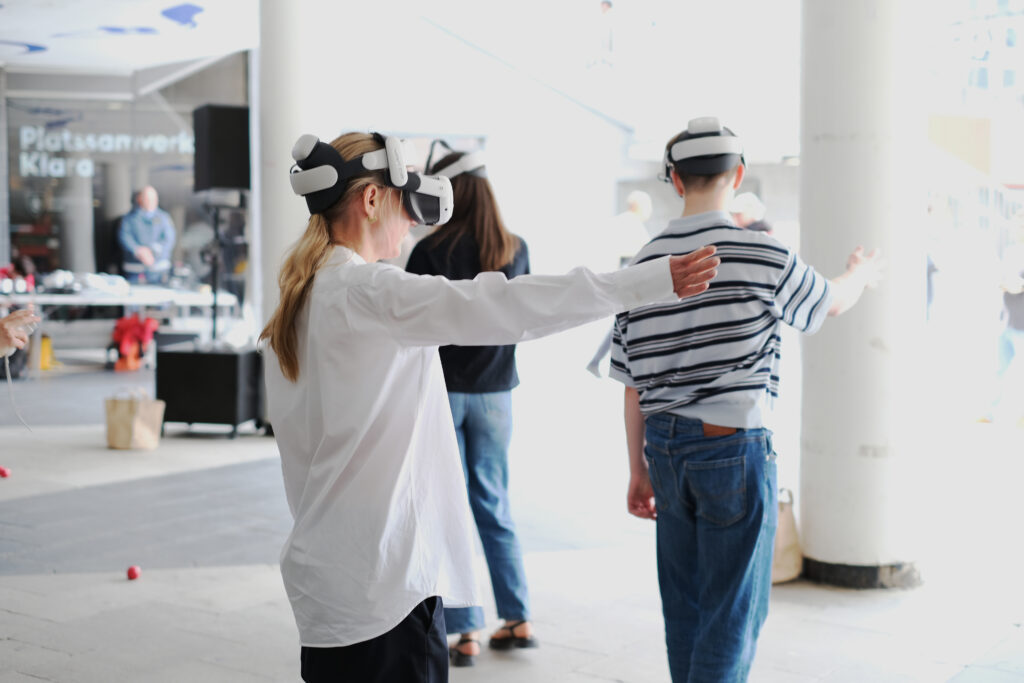 People wearing VR headsets stand with arms outstretched in a bright indoor space, engaging in a virtual reality activity. Some are facing away, and the area features white columns and blurred figures in the background.