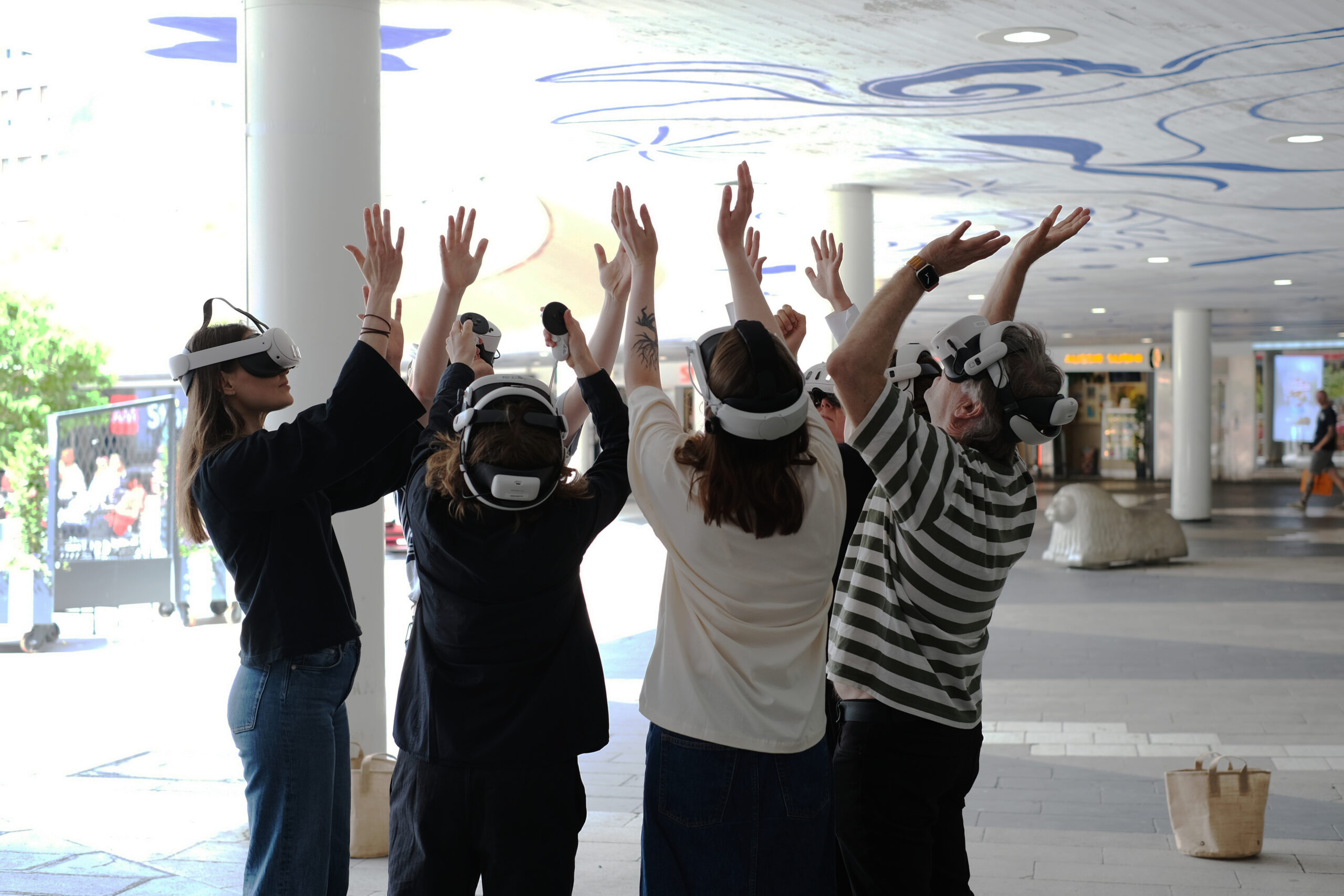 Four people wearing VR headsets stand outdoors with their arms raised, facing away from the camera, under a modern-looking, partially covered area.