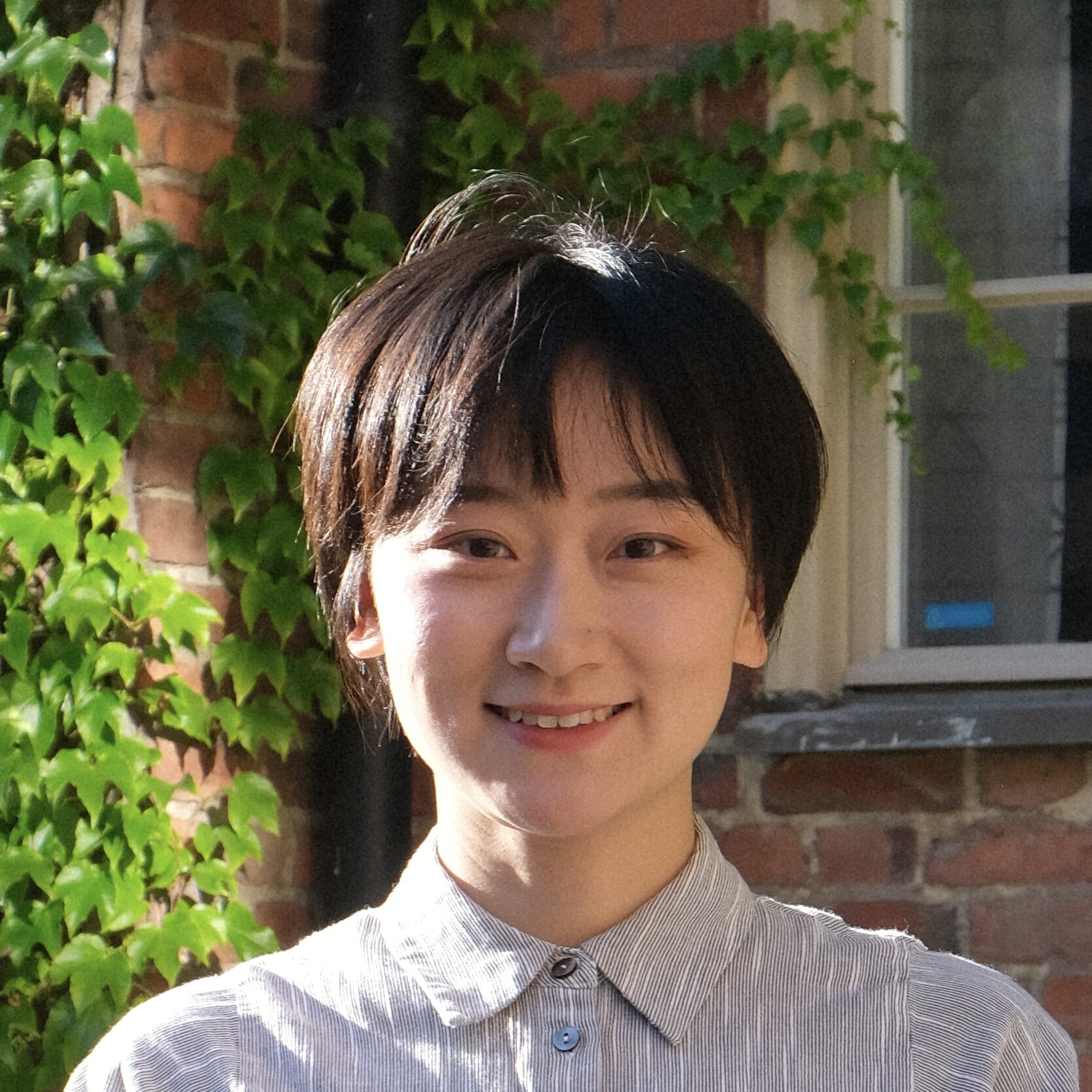 A person with short dark hair and a striped button-up shirt smiles outdoors, standing in front of a brick wall with green ivy and a window.