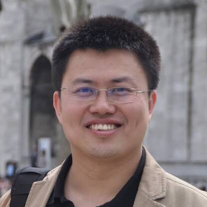 A man with short black hair and glasses smiles at the camera, wearing a beige jacket over a black shirt, with a blurred stone building in the background.