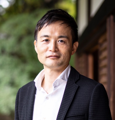 A man with short dark hair, wearing a white shirt and dark jacket, stands outdoors near a wooden building with greenery in the background.