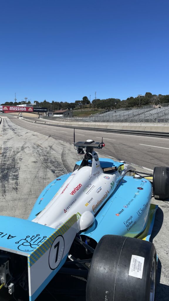 A blue and white open-wheel racing car with the number 1 is parked on a motorsport circuit under a clear blue sky, near empty stands and a Mission banner in the background.