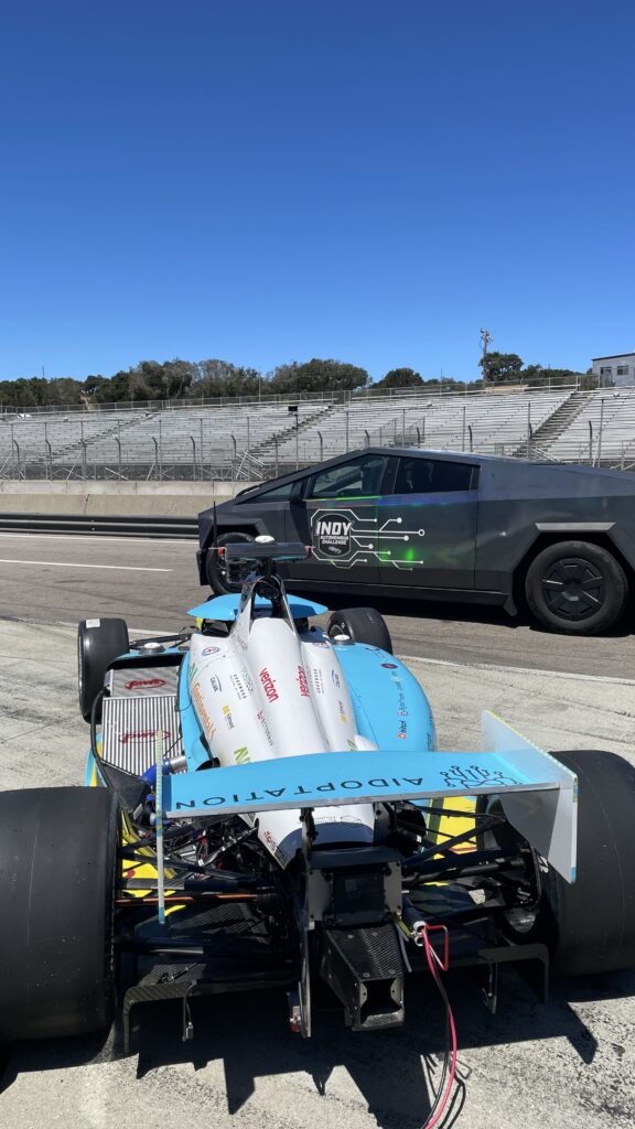 A blue and white formula racing car is parked on a track next to a black lorry with INDY branding. Empty grandstands and a clear blue sky are in the background.