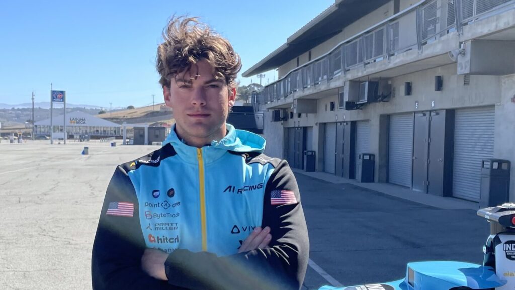 A person with wavy hair stands with arms folded in front of a blue and yellow racing car on a sunny day at a racetrack, with garages and empty grandstands in the background.