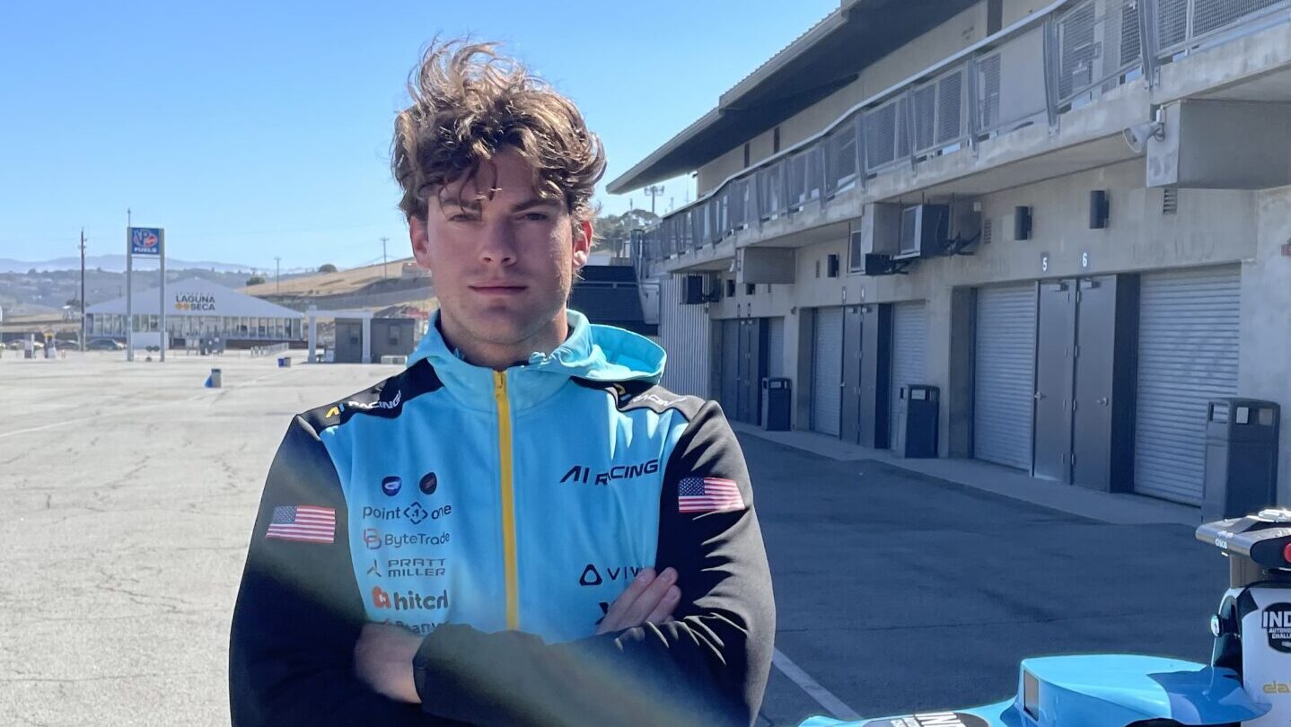 A person with wavy hair stands with arms folded in front of a blue and yellow racing car on a sunny day at a racetrack, with garages and empty grandstands in the background.