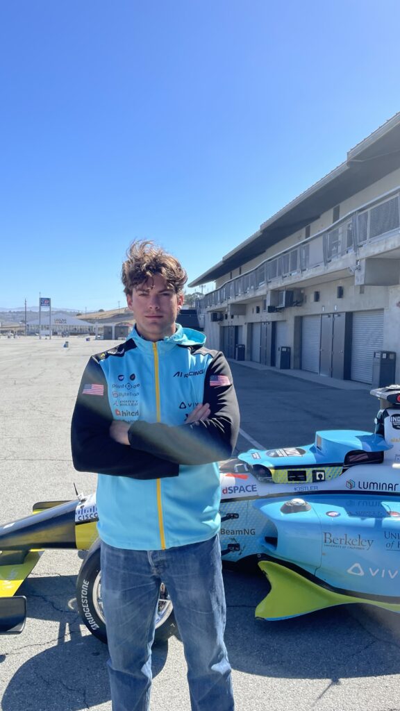 A person stands with arms folded in front of a blue and yellow racing car on a sunny day at a racetrack, with empty garages and clear blue sky in the background.