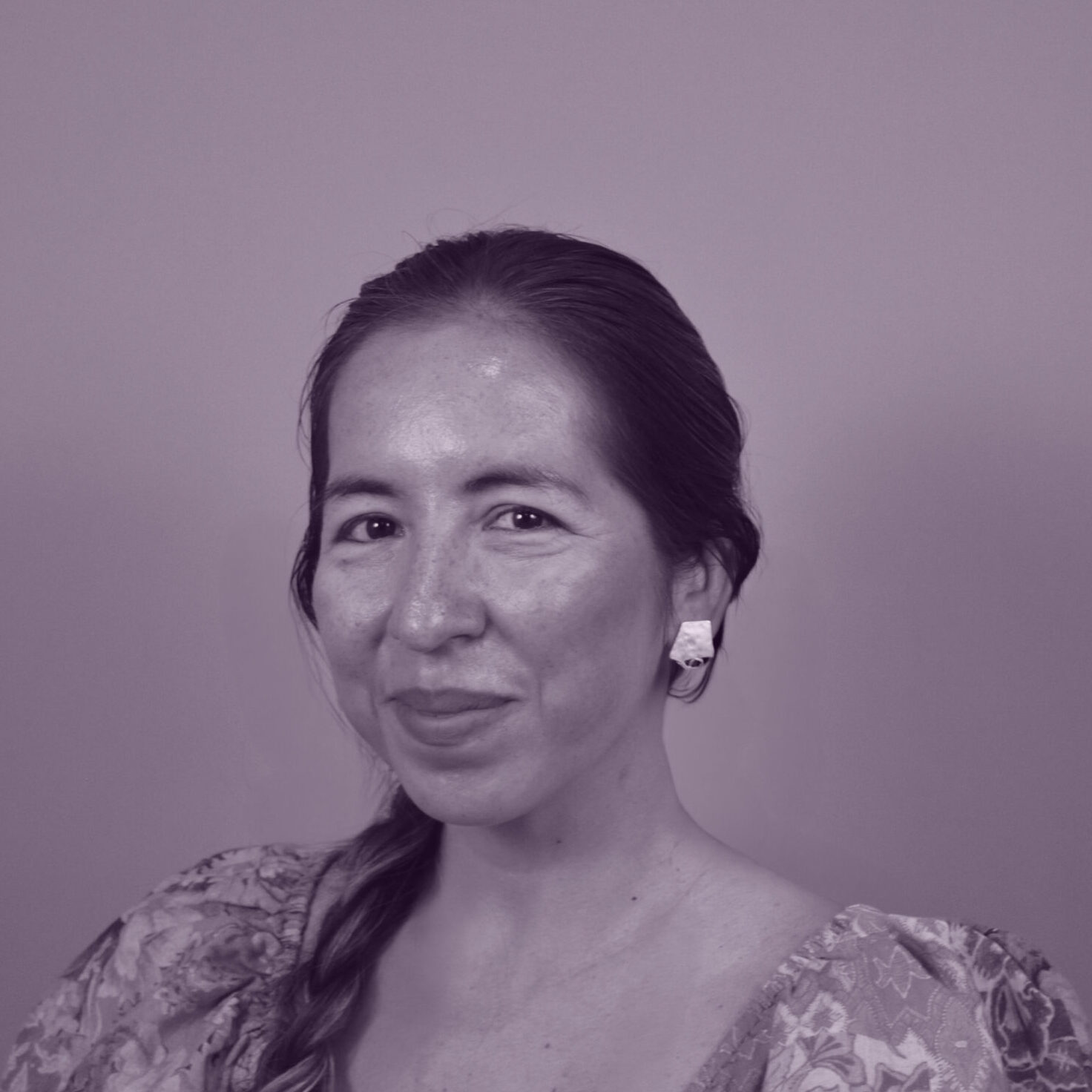 A woman with long hair in a plait, wearing a floral dress and earrings, smiles softly whilst posing against a plain background in a purple-tinted photograph.