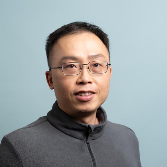 A man with short black hair and glasses wearing a grey fleece jacket stands in front of a plain light blue background, smiling slightly.