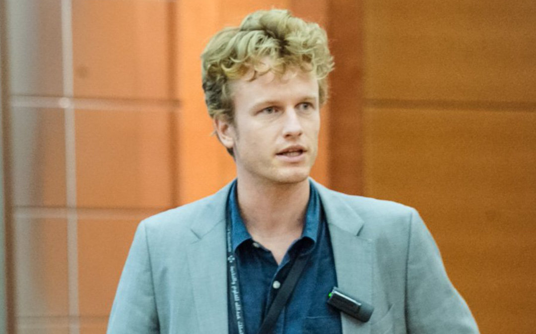 A man with blond curly hair, wearing a grey blazer over a dark shirt, stands indoors with a microphone clipped to his jacket.