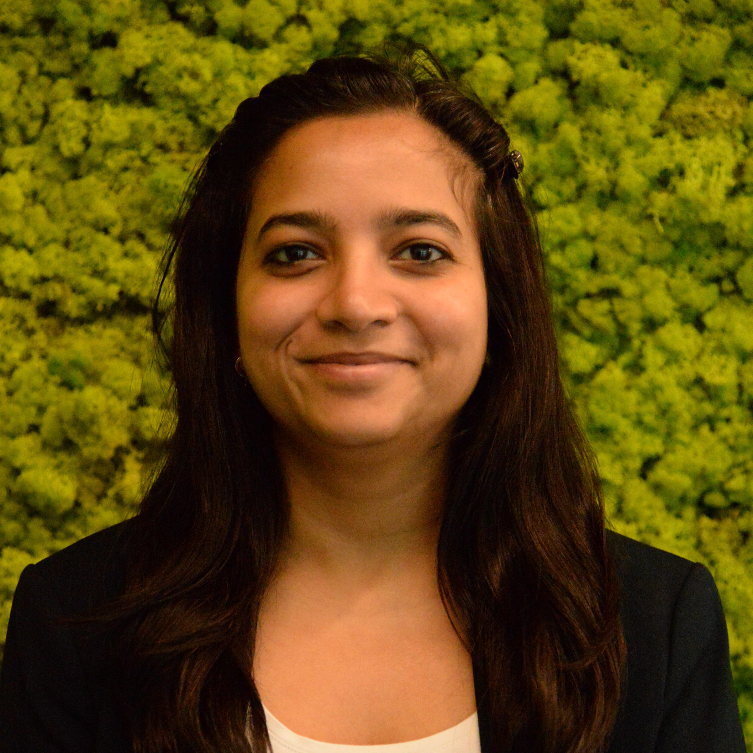 A woman with long dark hair, wearing a black blazer and white top, smiles in front of a green, leafy backdrop.