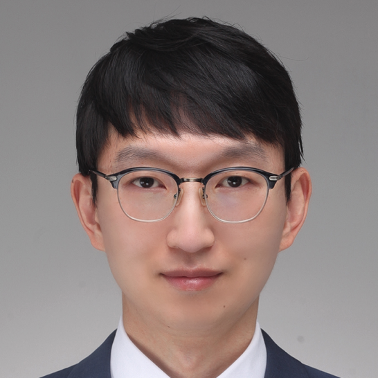 A young man with short black hair, wearing round glasses, a white dress shirt, and a blue suit jacket, poses against a plain grey background.