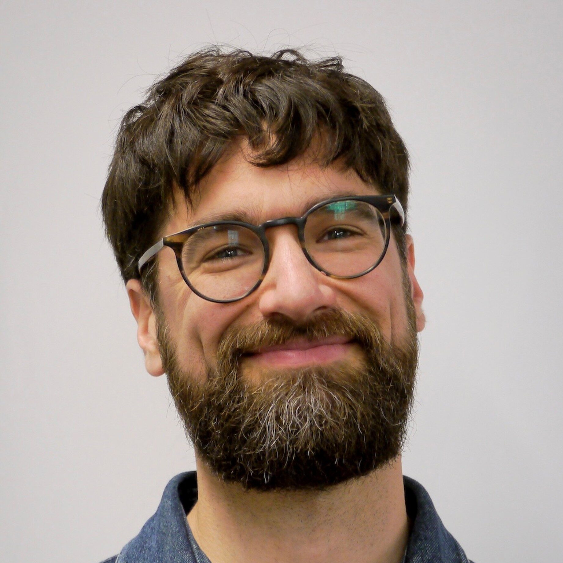 A man with brown hair, a beard, and glasses smiles at the camera. He is wearing a denim collared jacket over a white shirt. The background is plain and light-coloured.