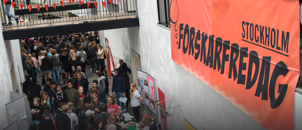 A large crowd gathers indoors beneath an orange banner reading Stockholm ForskarFredag. People of all ages are interacting at tables, and triangular bunting decorations hang above.