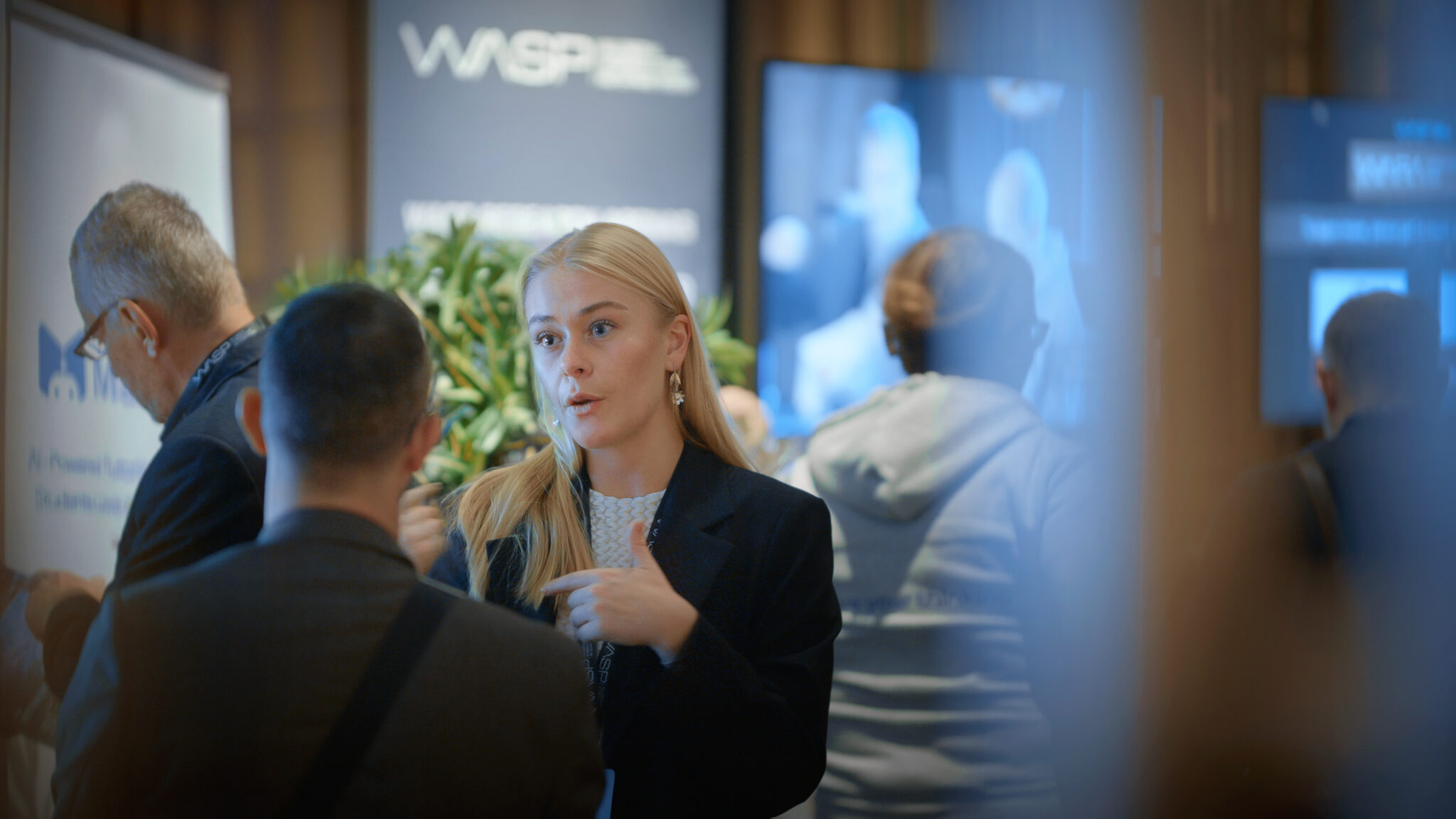 A woman in a black jacket speaks to a man at a busy indoor event, with blurred people and informational signs in the background.
