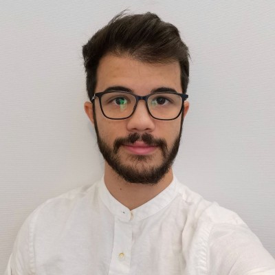A young man with short brown hair, a beard, and glasses is wearing a white shirt and looking at the camera against a plain light-coloured background.