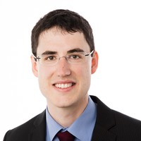 A young man with short dark hair, wearing glasses, a dark suit, blue shirt, and burgundy tie, smiles at the camera against a plain white background.
