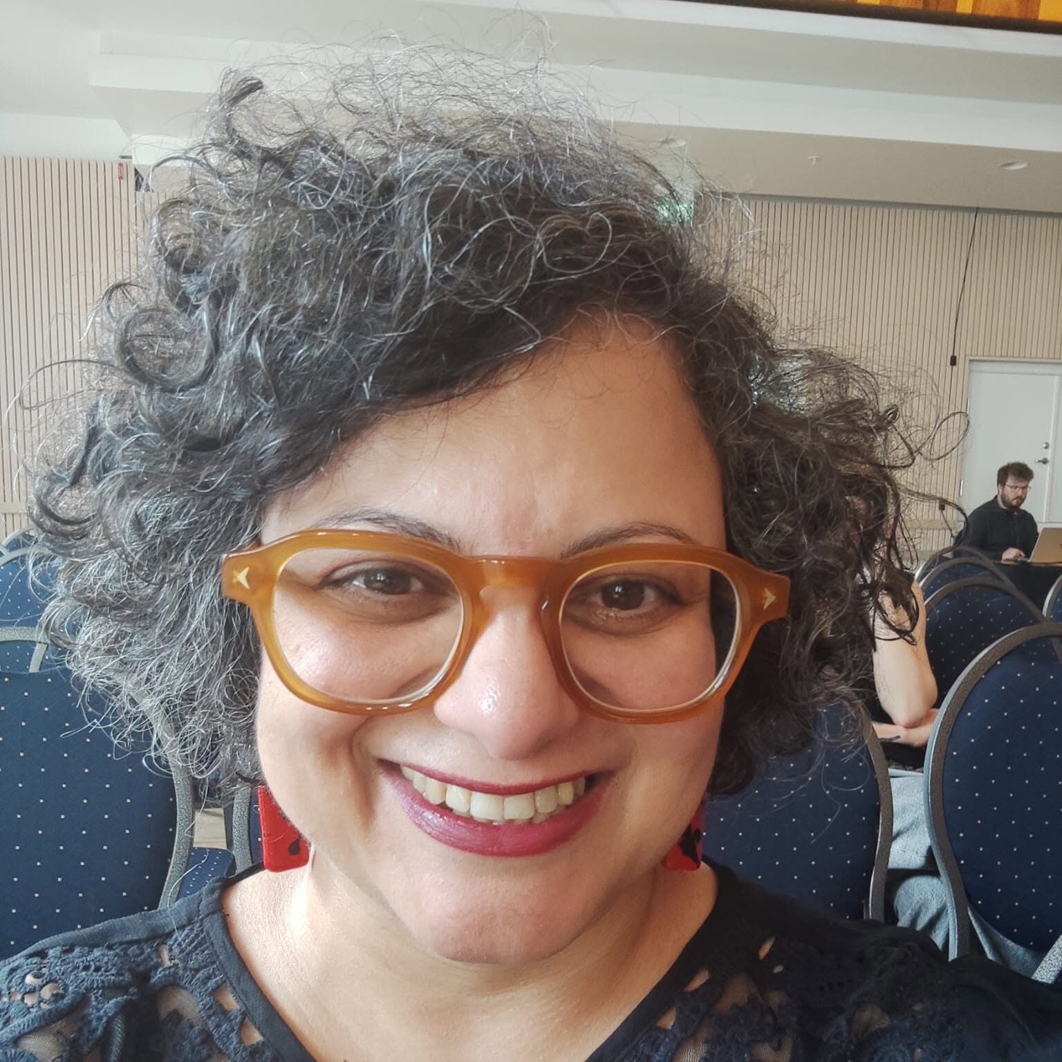 A person with curly grey hair and brown glasses smiles at the camera, wearing a black lace top. Blue chairs and other people are visible in the background.
