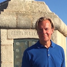A man in a blue polo shirt stands in front of a stone structure with a plaque that reads GARITA DE LA. The sky is clear and blue in the background.