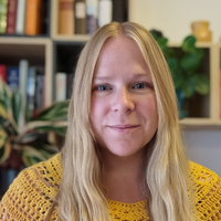 A woman with long blonde hair wearing a yellow crochet top sits indoors in front of bookshelves and plants, smiling softly at the camera.