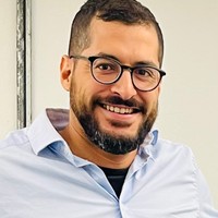A man with short hair, a beard, and glasses smiles at the camera. He is wearing a light blue collared shirt over a black shirt and stands against a plain, light-coloured background.