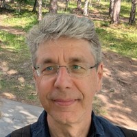 A man with grey hair, glasses, and a slight smile stands outdoors in a sunlit wooded area with green grass and trees in the background.