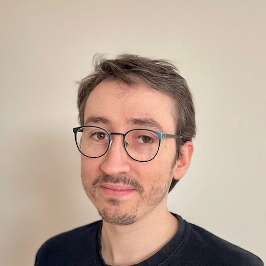 A person with short brown hair, glasses, and light facial hair wears a black shirt and looks at the camera, standing in front of a plain light-coloured background.
