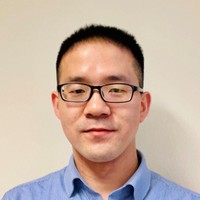 A man with short black hair and glasses, wearing a light blue collared shirt, stands in front of a plain off-white background, smiling slightly at the camera.