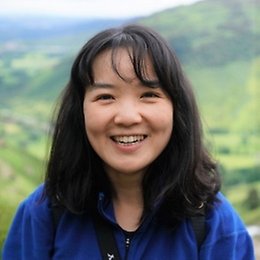 A woman with medium-length black hair wearing a blue jacket smiles at the camera, standing outdoors with green hills and a blurred landscape in the background.