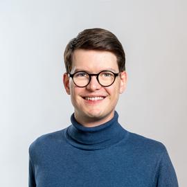 A smiling person with short brown hair, wearing round glasses and a blue polo-neck jumper, stands against a plain light background.