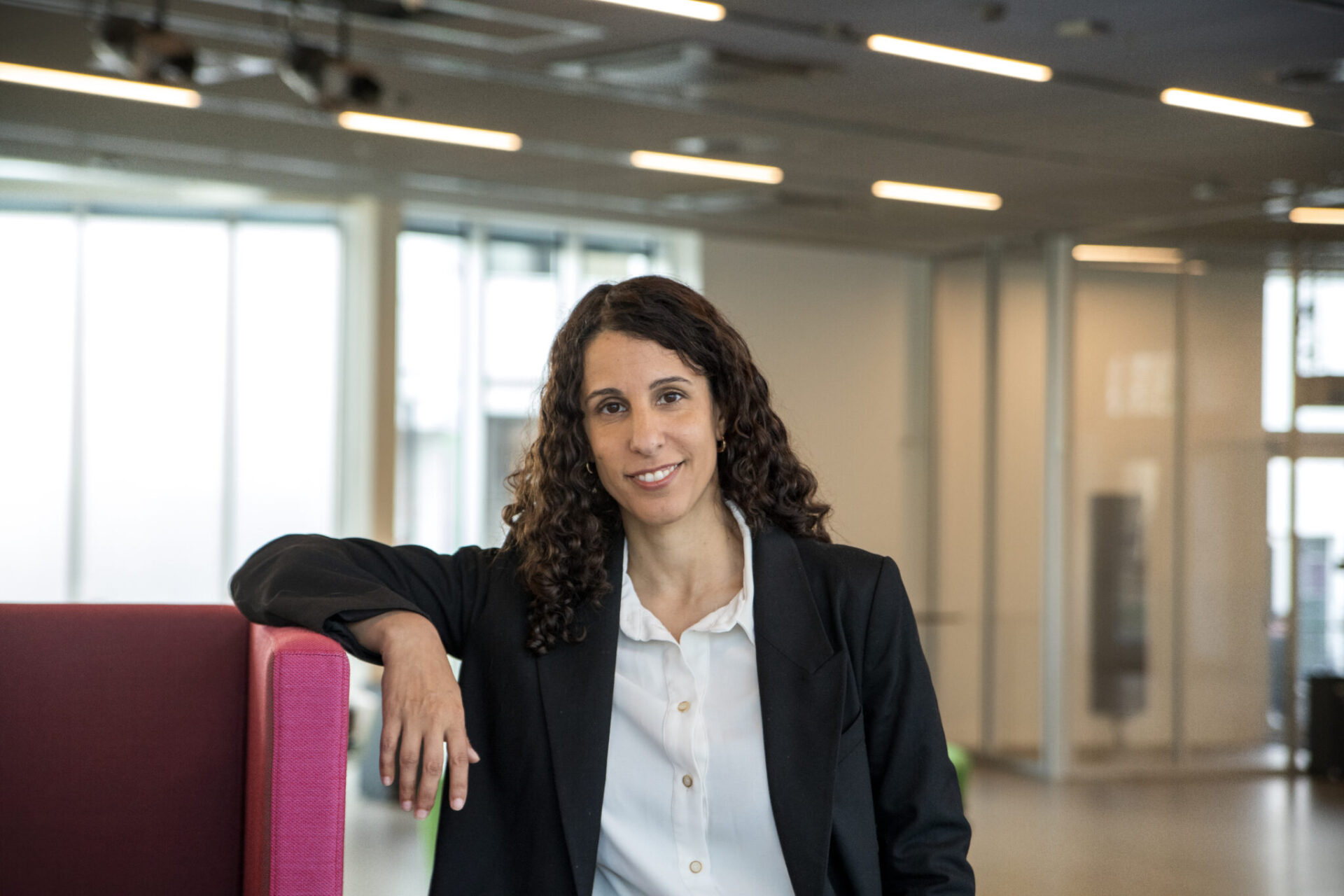 A woman with curly hair in a black blazer and white shirt smiles, leaning on a pink chair in a bright, modern office space with large windows in the background.