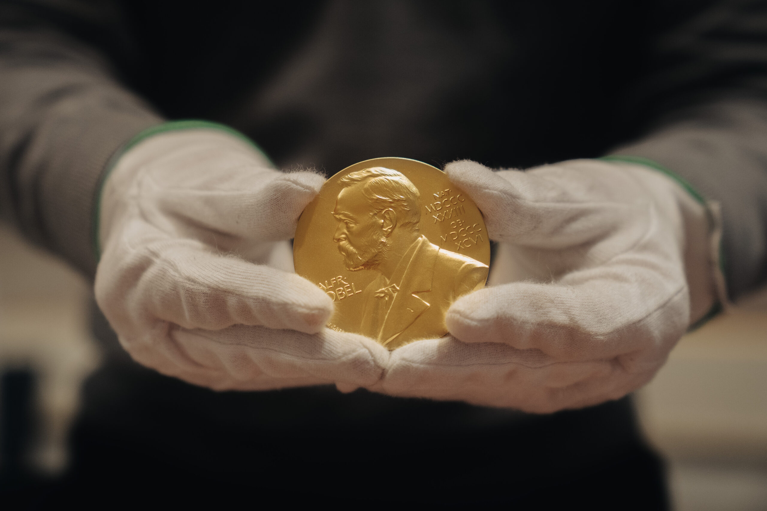 A person wearing white gloves holds a Nobel Prize medal, featuring a profile of Alfred Nobel engraved on its surface.