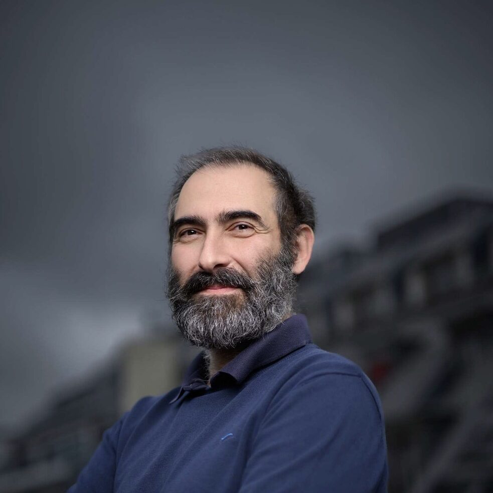 A man with a beard and moustache, wearing a blue long-sleeve shirt, stands outdoors with his arms crossed and smiles slightly. The background is an overcast sky and a blurred building.