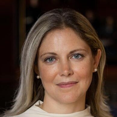 A woman with long blonde hair and blue eyes, wearing a cream-coloured top, looks at the camera with a slight smile. The background is softly blurred.