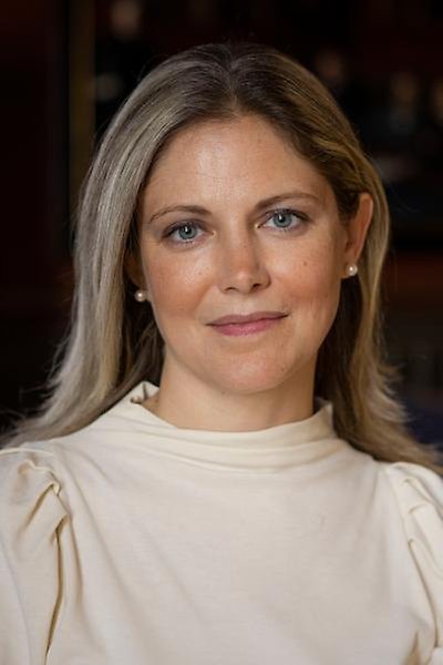 A woman with long blonde hair and blue eyes, wearing a cream-coloured top, looks at the camera with a slight smile. The background is softly blurred.