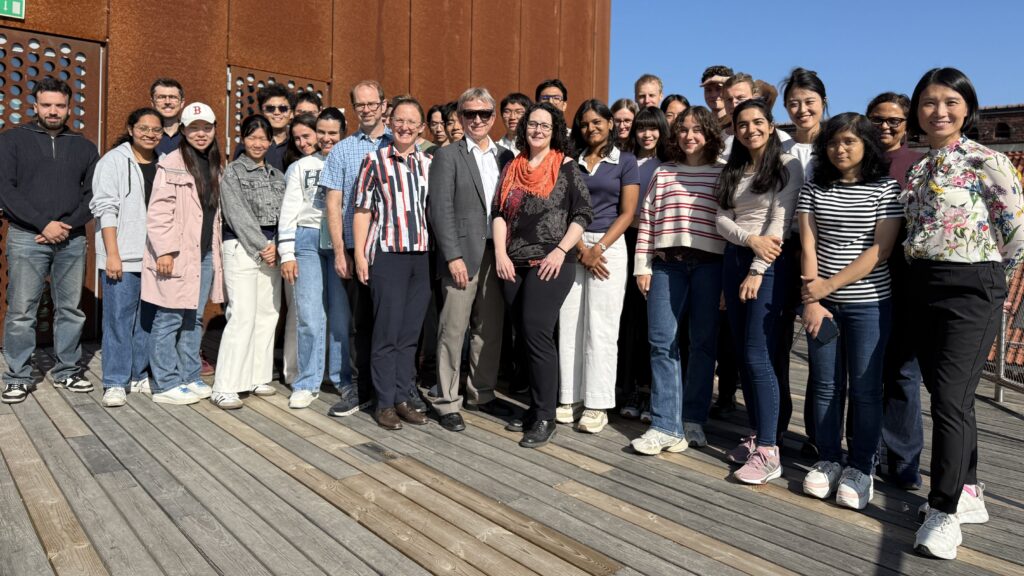 A group of about 25 people stand together on a wooden decking in sunlight, posing and smiling for a group photo. A rust-coloured metal wall and bright blue sky are in the background.