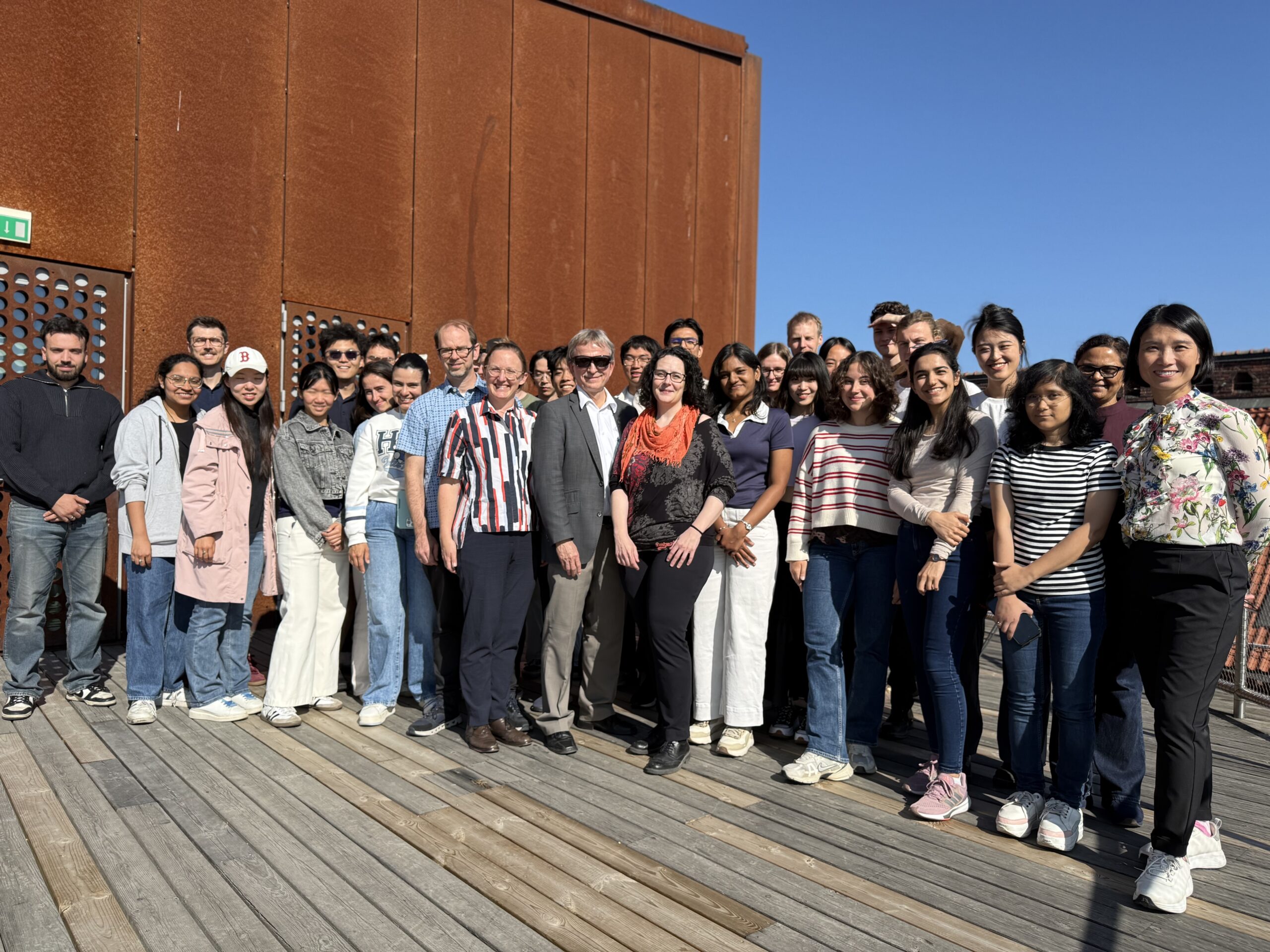 A group of about 25 people stand together on a wooden decking in sunlight, posing and smiling for a group photo. A rust-coloured metal wall and bright blue sky are in the background.