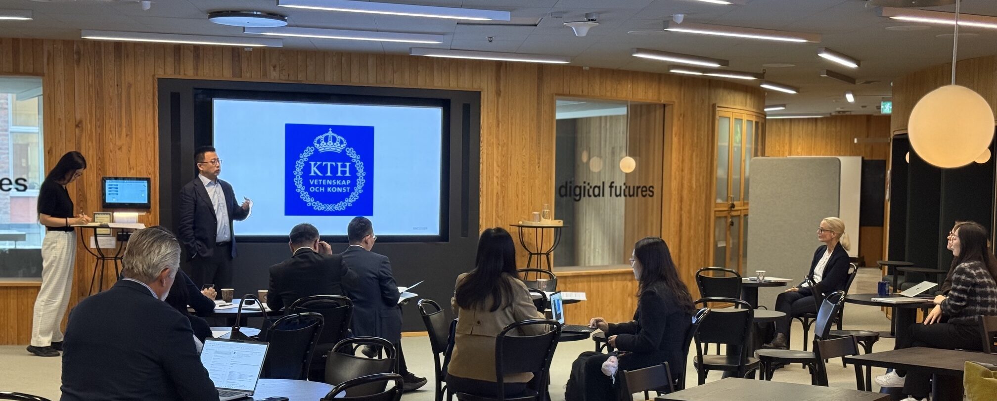 A small group of people sit spaced apart in a modern conference room. A man presents at the front with a KTH logo on the screen. Some attendees use laptops.
