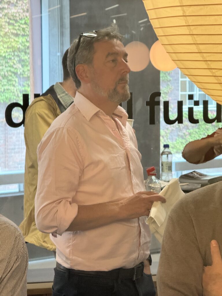 A man in a light pink shirt holds a water bottle and serviette, standing indoors near a window with text in the background. Other people are partially visible around him.