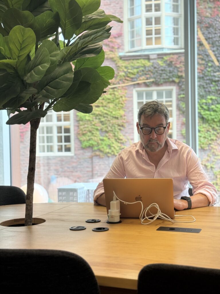 A man in glasses and a pink shirt works on a laptop at a wooden table in a modern office with large windows and a potted tree.