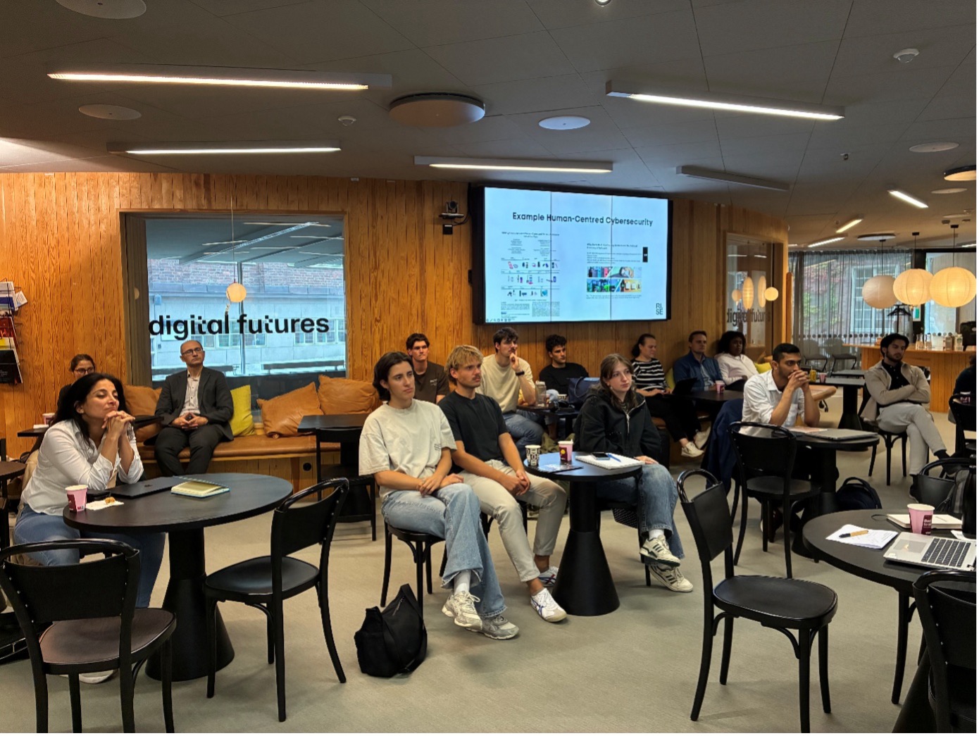 A group of people sit in a modern meeting room, attentively watching a presentation on a screen labelled Human-Centred Cybersecurity. The space features wood walls, glass, and digital futures signage.