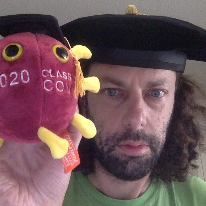 A person wearing a graduation cap holds a soft toy shaped like a virus, also wearing a cap, with 2020 Class Cov stitched on it.