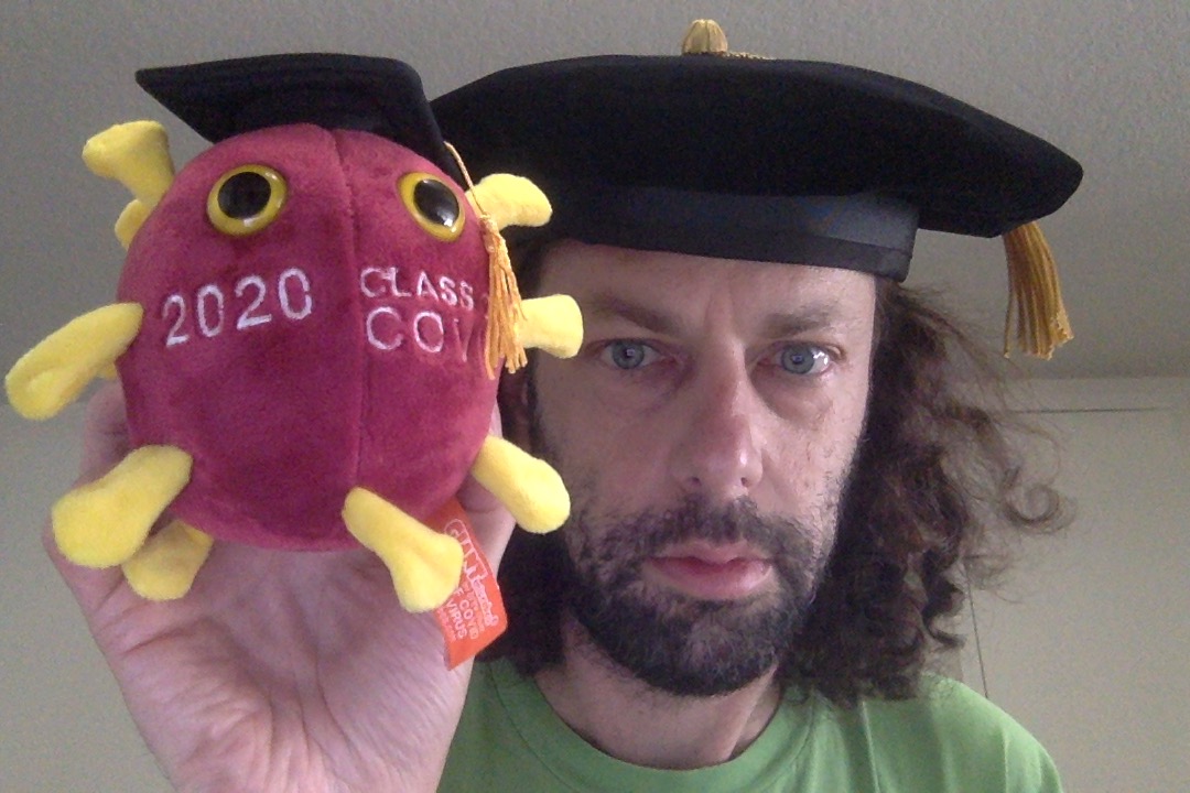 A person wearing a graduation cap holds a soft toy shaped like a virus, also wearing a cap, with 2020 Class Cov stitched on it.