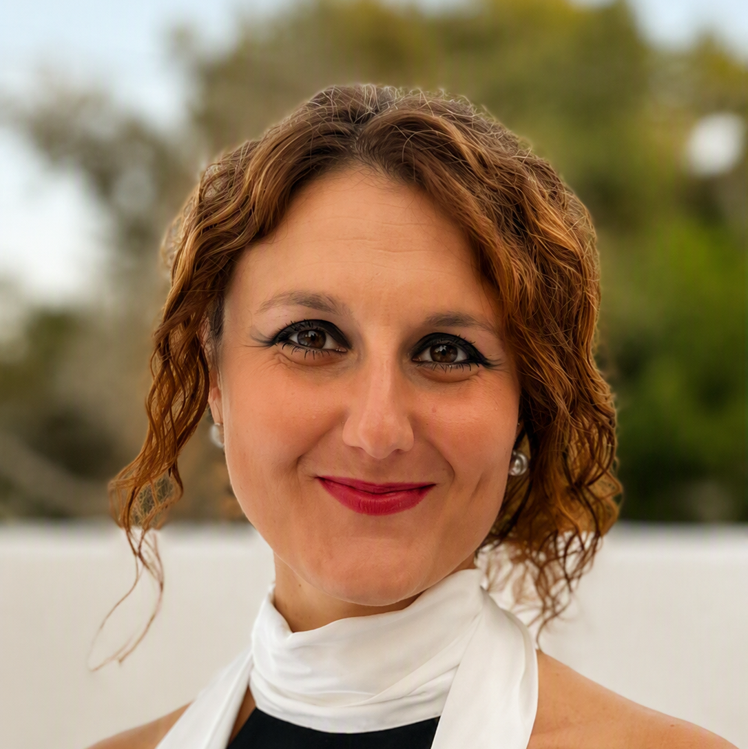 A woman with curly brown hair smiles at the camera, wearing a sleeveless black top and a white scarf. The blurred outdoor background of green trees adds a natural touch to this portrait of Liviana Palmisano.