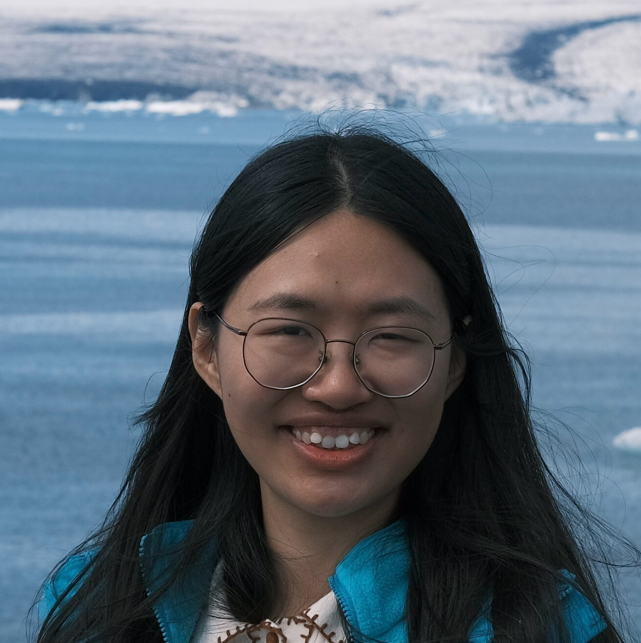 A woman with long black hair and glasses smiles outdoors, standing in front of a body of water with snow-covered mountains in the background. She wears a blue jacket and a white top.