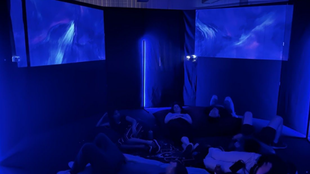 People lie on the floor in a dark room illuminated by blue light, watching swirling blue and purple visuals projected onto screens on the walls. The scene has a calm, immersive, and meditative atmosphere.