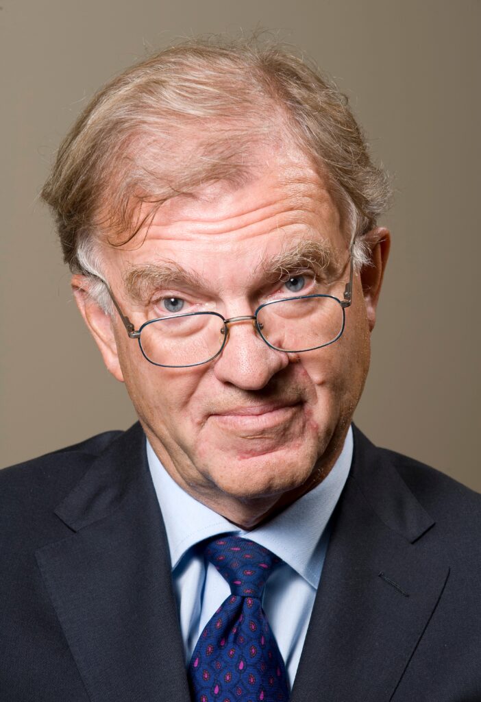 An older man in a suit and patterned tie looks directly at the camera with a slight smile. He wears glasses and has thinning, light brown hair. The background is plain and neutral.
