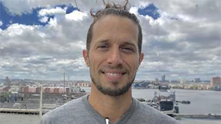A man with long hair tied back smiles at the camera with a cityscape and waterfront visible behind him under a partly cloudy sky.