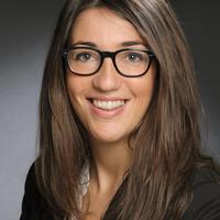 Smiling woman with long brown hair and glasses, wearing a black blazer and white top, posed in front of a dark background.
