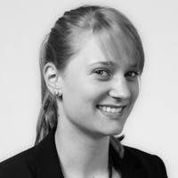 Black and white portrait of a woman with fair hair pulled back, wearing a dark blazer and small earrings, smiling at the camera against a plain background.